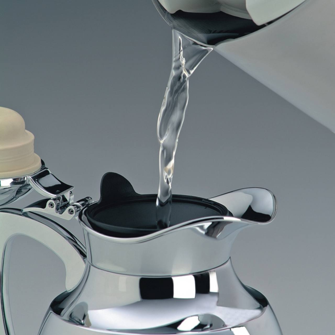 Ein Wasserkocher aus Metall wird mit Wasser von einem Wasserhahn befüllt. Der Wasserstrahl fließt direkt in die Öffnung des Kocherdeckels. Die Umgebung wirkt modern und aufgeräumt.