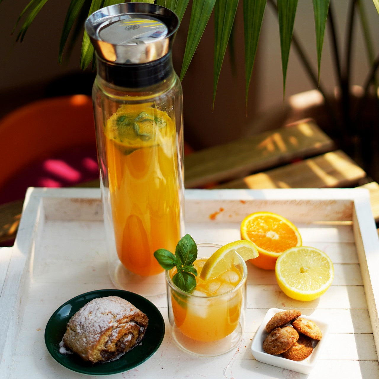 Auf einem Tablett steht ein Glas mit oranger Flüssigkeit, garniert mit Limettenscheiben und Minzblättern. Daneben liegen geschnittene Orangen und Zitronen sowie ein Schälchen mit Salz und Nüssen. Im Hintergrund sind unscharfe Pflanzen erkennbar.