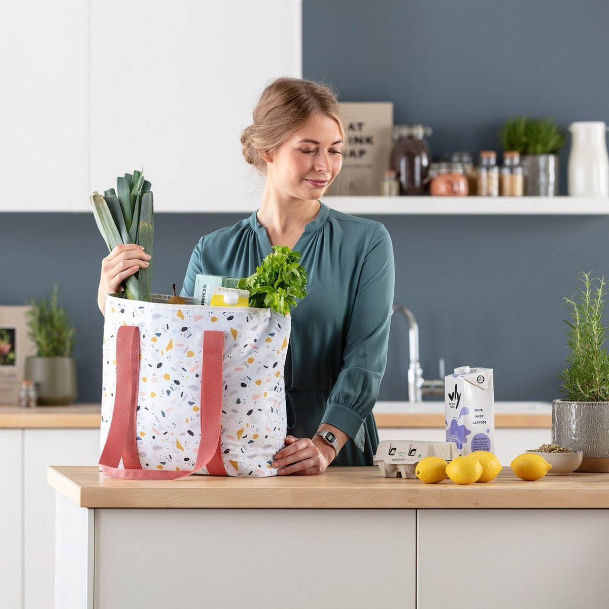 Eine Frau steht in einer modernen Küche und räumt eine Einkaufstasche voller frischer Lebensmittel aus. Auf der Arbeitsplatte liegen Zitronen, ein Eierkarton und eine Milchtüte. Sie trägt ein langärmeliges, grünes Kleid und lächelt.