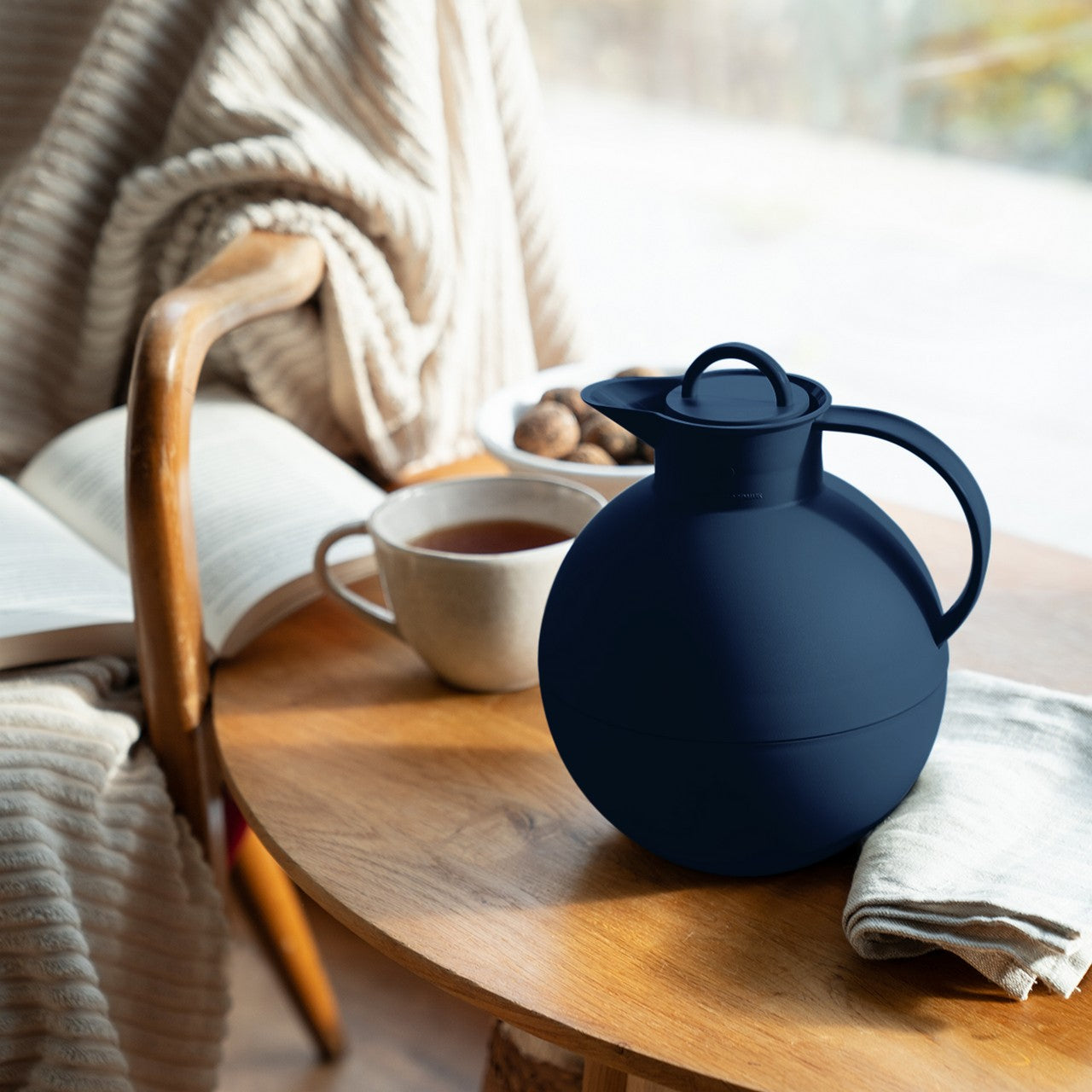 Ein runder Holztisch zeigt eine blaue Kanne, eine Tasse Tee und ein gefaltetes Tuch. Ein gepolsterter Stuhl mit beigefarbener Decke trägt ein offenes Buch. Im Hintergrund erhellt ein großes Fenster die Szene.