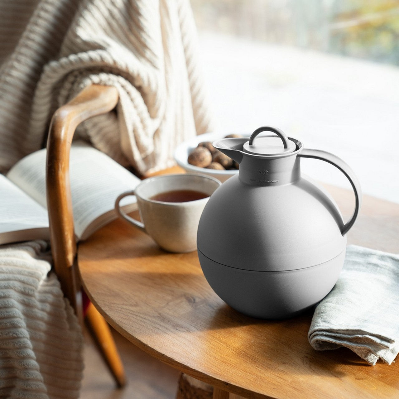 Auf einem Tisch vor einem Fenster stehen eine graue Thermoskanne, eine Tasse Tee und ein gefaltetes Tuch. Daneben auf einem Stuhl liegen eine Decke und ein aufgeschlagenes Buch. Im Hintergrund sind Nüsse auf einem Teller.