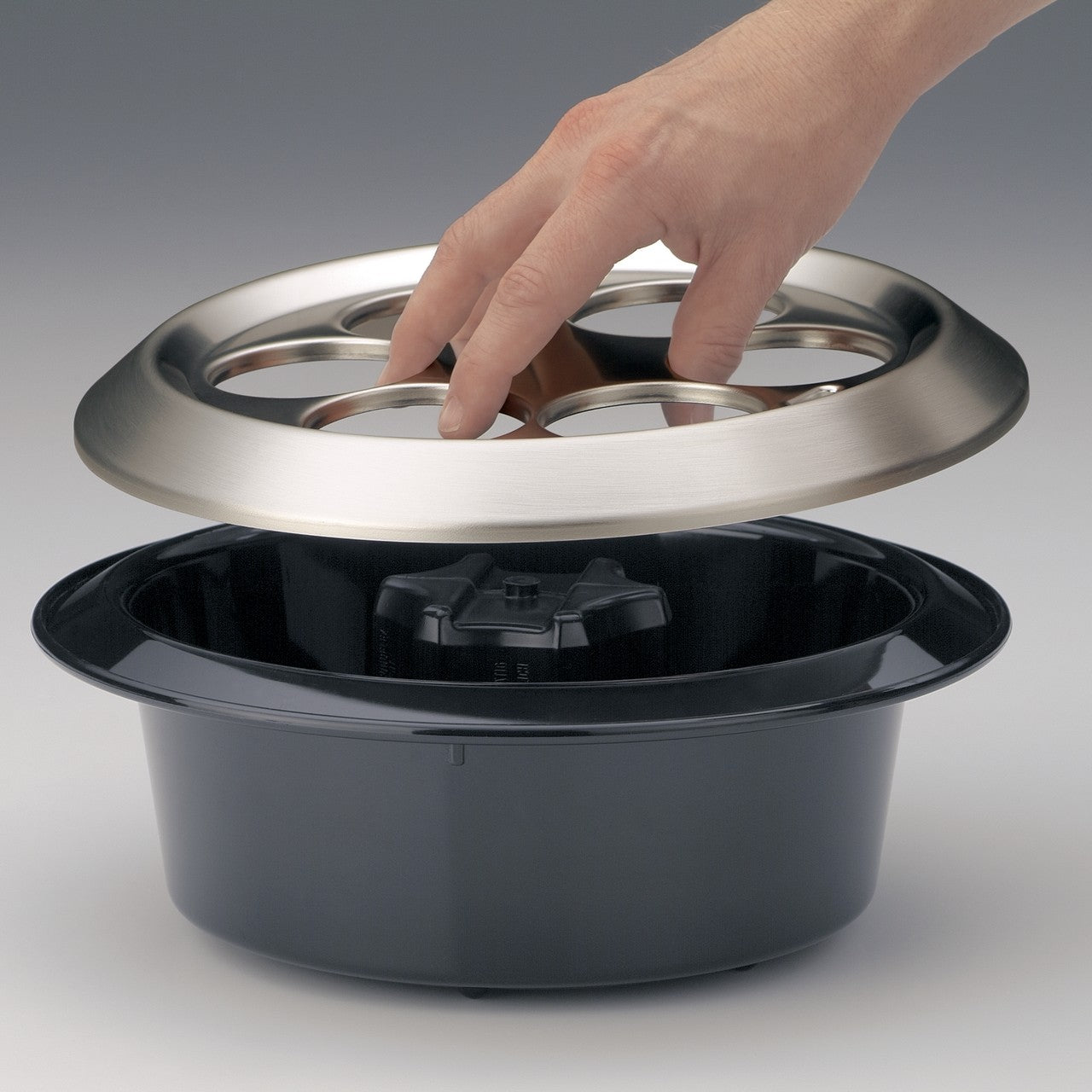 Ein Topf mit einem metallischen Deckel wird geöffnet. Eine Hand hebt den Deckel leicht an. Der Topf ist dunkel und die Szene hat einen neutralen Hintergrund.