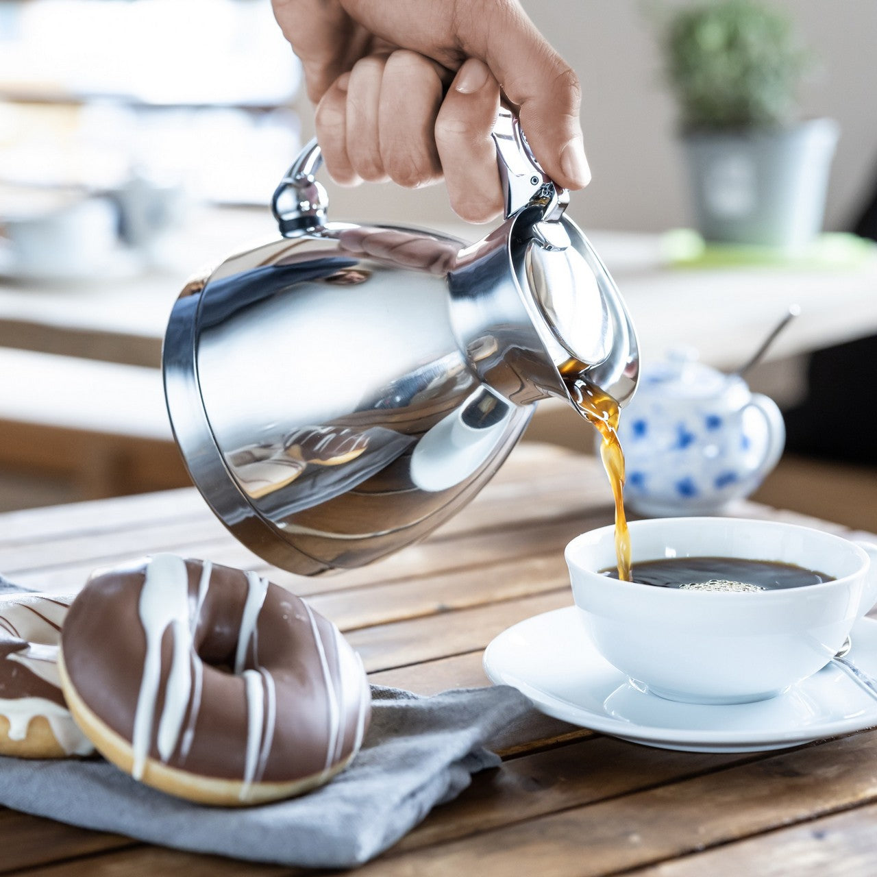Ein silberner Teekessel gießt Tee in eine weiße Tasse, die auf einem Holztisch steht. Neben der Tasse liegt ein Gebäckstück mit Zuckerglasur. Der Hintergrund zeigt einen gemusterten Zuckertopf und weitere verschwommene Gegenstände.