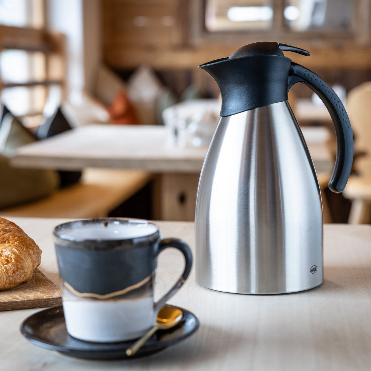 Ein silberner Isolierkrug mit schwarzem Griff steht auf einem Holztisch. Daneben sind eine schwarze Tasse mit Goldlöffel und ein Croissant zu sehen. Der Hintergrund zeigt einen gemütlichen Raum mit Holzmöbeln.