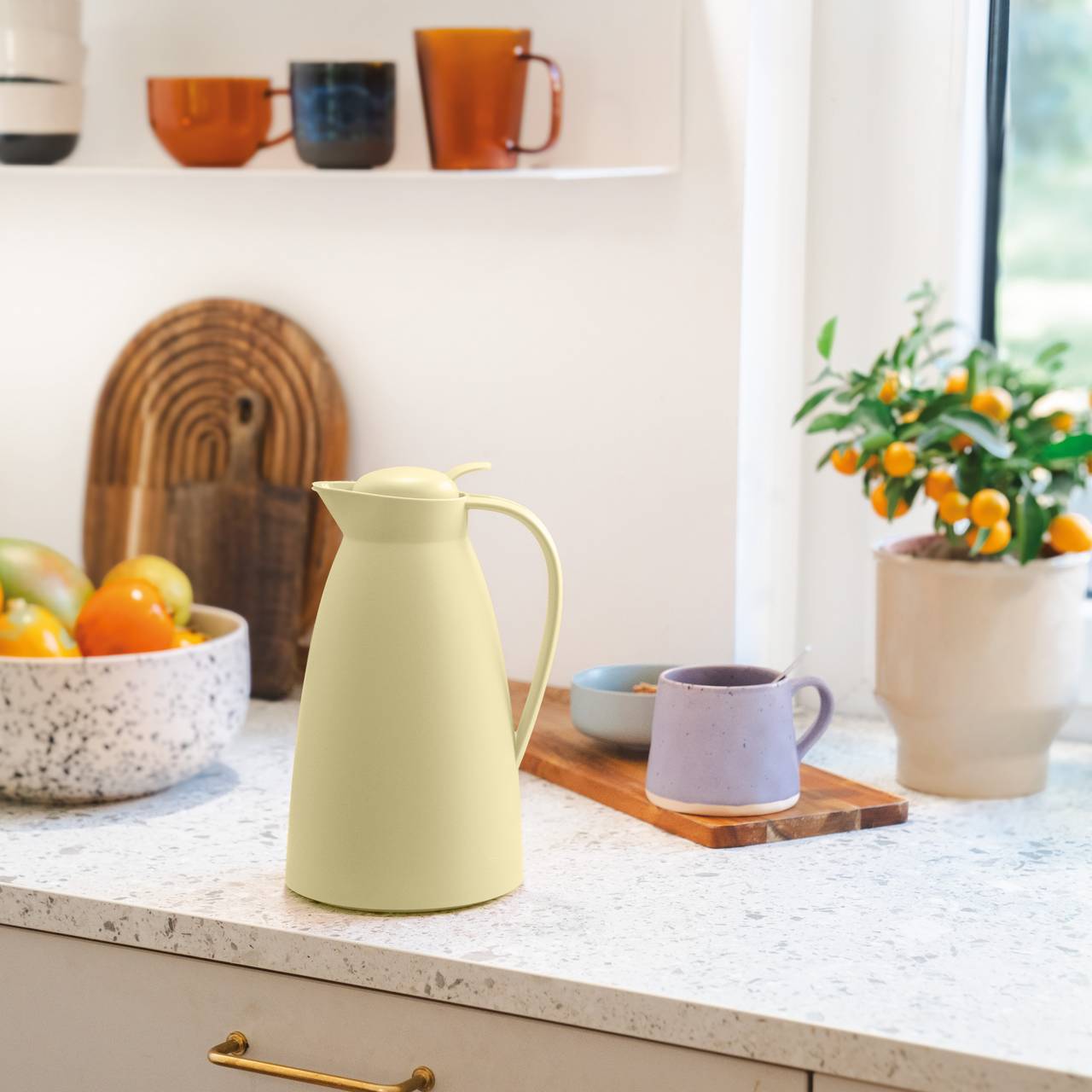 Auf der Küchenarbeitsplatte steht eine helle Thermoskanne neben einer Schale mit buntem Obst und einer Tasse auf einem Holzbrett. Im Hintergrund sind weitere Tassen und Pflanzen zu sehen. Durch das Fenster fällt Tageslicht in den Raum.