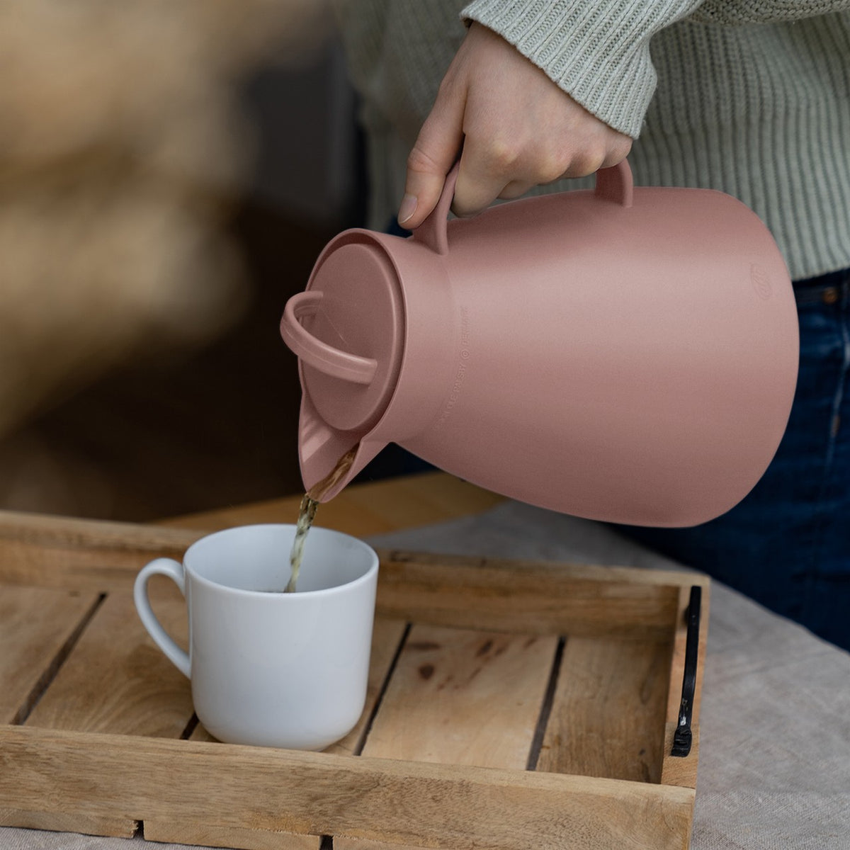 Eine Person gießt aus einer rosafarbenen Thermoskanne eine Flüssigkeit in eine weiße Tasse, die auf einem hölzernen Tablett steht. Die Person trägt einen hellgrauen Pullover.