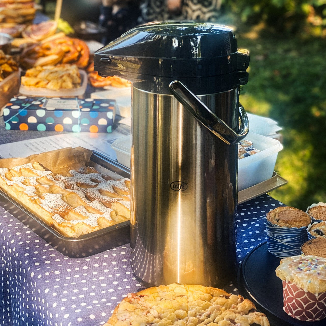 Auf dem Bild ist ein silberner Pump-Thermosbehälter zu sehen, der auf einem Tisch steht. Um ihn herum sind viele verschiedene Gebäckstücke und Kekse auf Tabletts präsentiert. Die Umgebung hat eine warme, einladende Atmosphäre.
