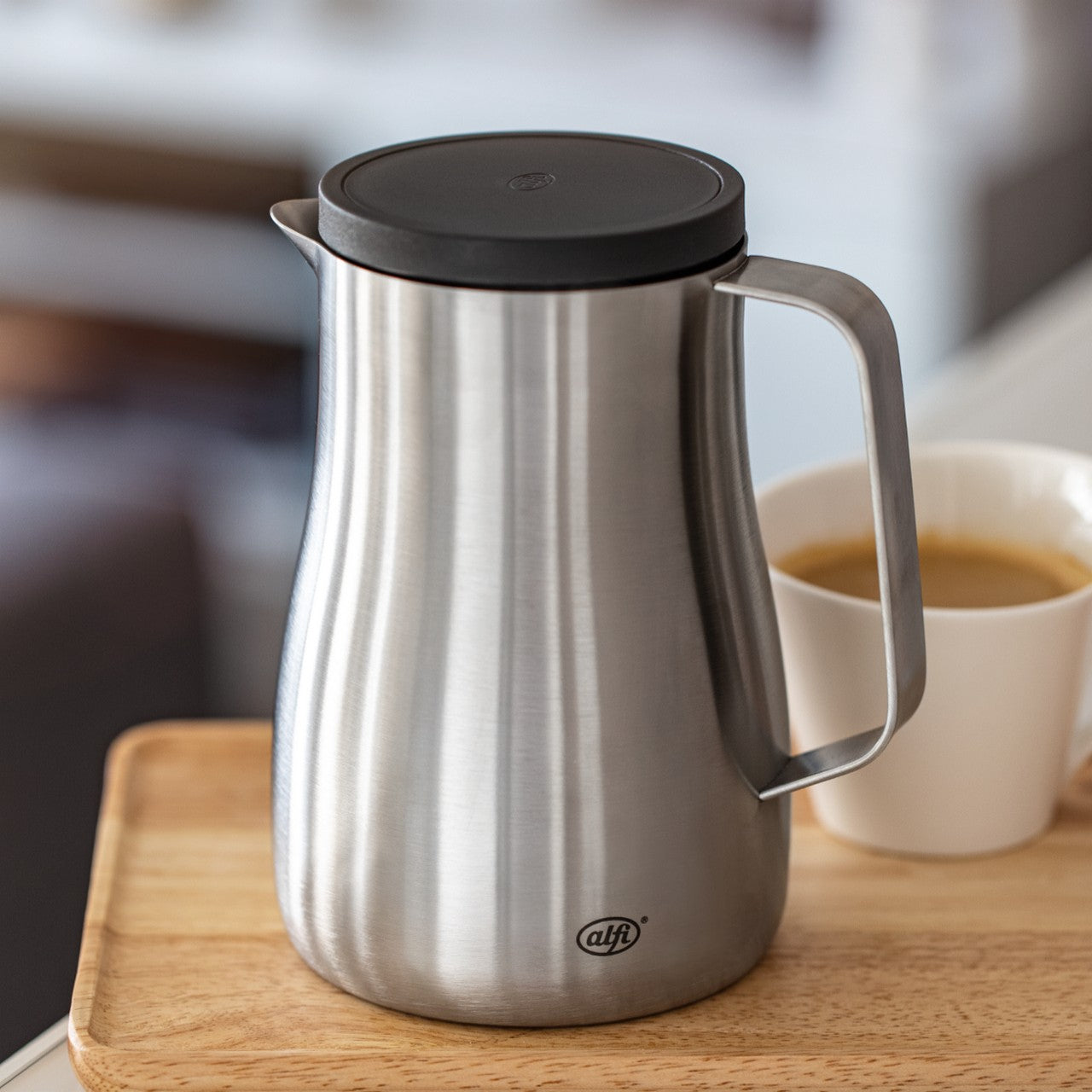 Auf dem Bild steht eine moderne, silberne Thermoskanne auf einem hölzernen Tablett. Daneben befindet sich eine weiße Tasse mit Kaffee. Der Hintergrund wirkt leicht verschwommen und vermittelt den Eindruck eines gemütlichen Wohnbereichs.