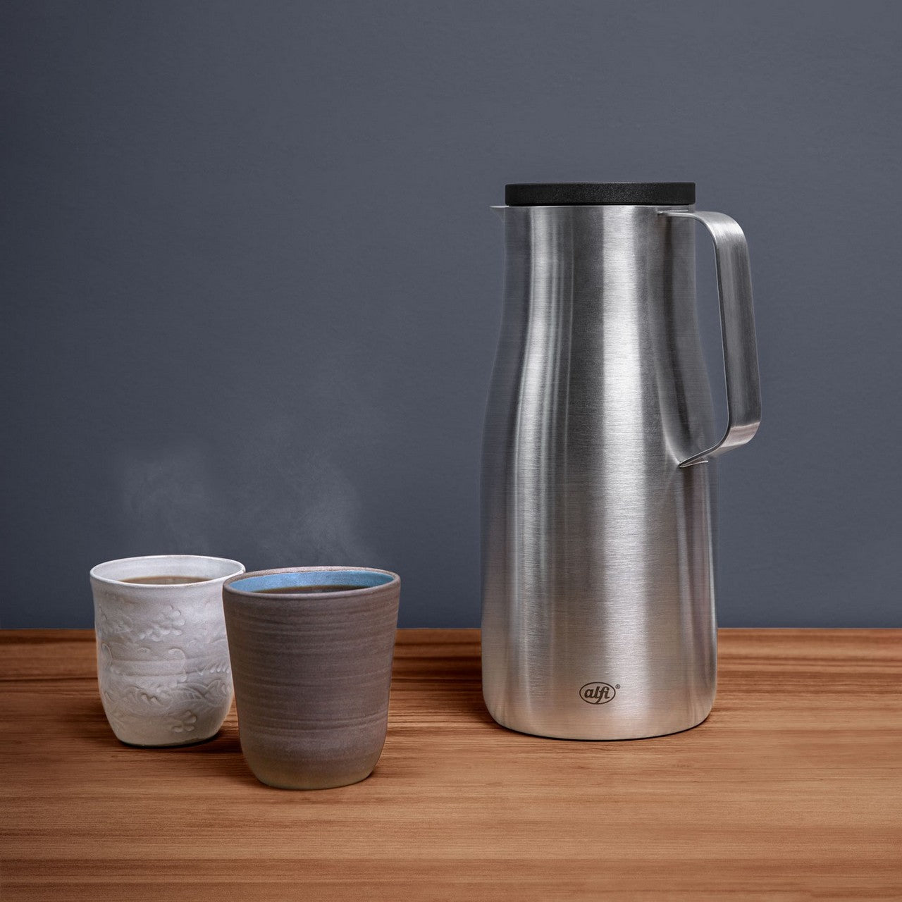 Auf einem Holztisch stehen eine silberne Thermoskanne aus Edelstahl und zwei Tassen, aus denen Dampf aufsteigt. Die Tassen sind schlicht, eine ist weiß und die andere grau. Der Hintergrund ist in einem schlichten Grauton gehalten.