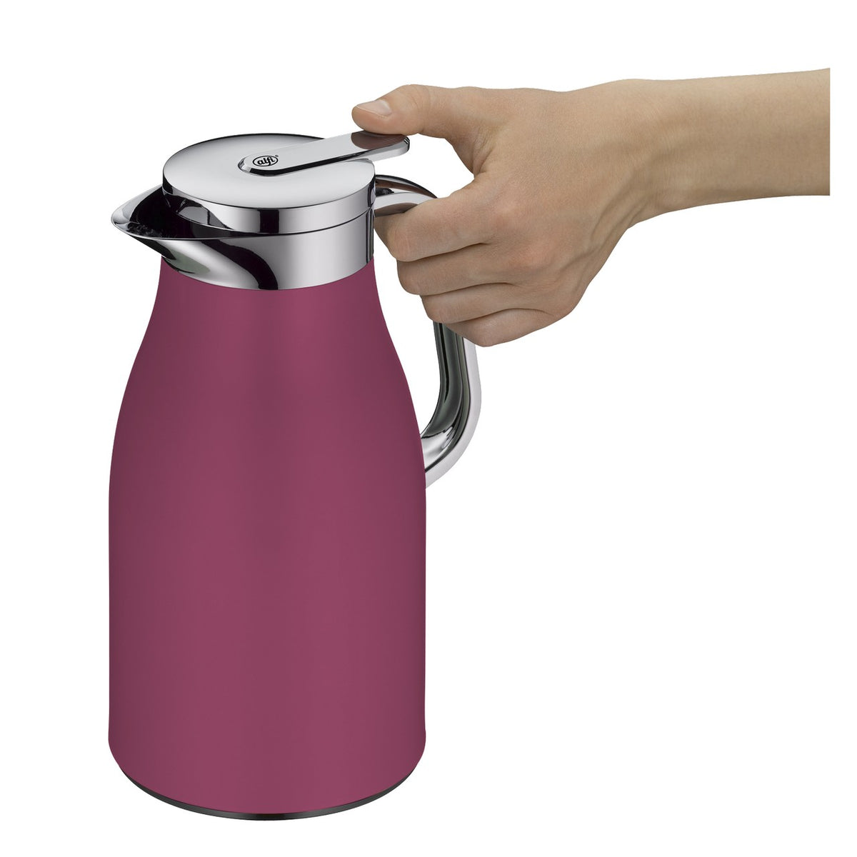 Auf einem Tisch steht eine rosa Thermoskanne neben einem Glas mit bunten Blumen und einer brennenden Kerze. Daneben liegen ein Croissant auf einem Teller, Bücher und eine Strickdecke. Im Hintergrund ist unscharf eine grüne Kulisse zu sehen.