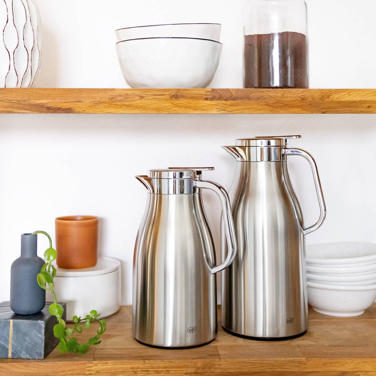 Zwei silberne Thermoskannen stehen auf einem Holzregal. Neben ihnen sind eine kleine graue Vase mit Ranke und ein orangefarbener Becher. Oben befinden sich weiße Schalen und ein Glas mit Kaffeepulver.