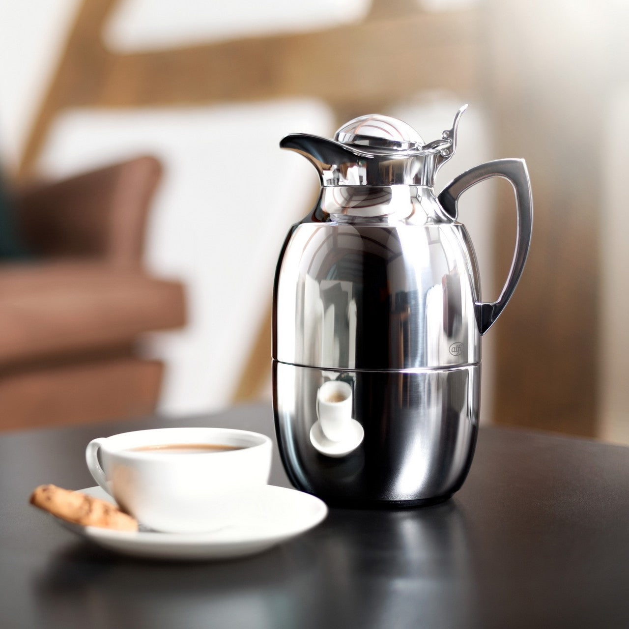 Eine glänzende Metall-Thermoskanne steht auf einem Tisch neben einer Kaffeetasse mit Milchschaum und einem Keks. Im Hintergrund ist eine verschwommene, gemütliche Sitzgelegenheit zu erkennen.
