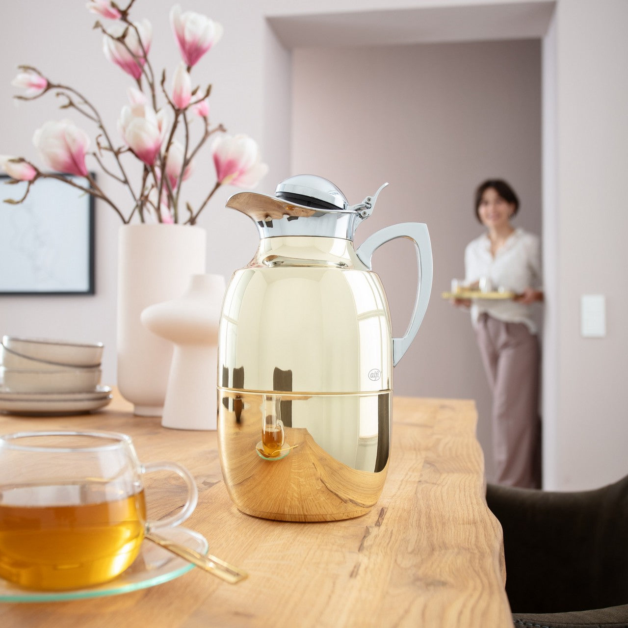 In einem stilvoll eingerichteten Raum steht eine glänzende goldene Thermoskanne auf einem Holztisch, neben einer Tasse Tee. Im Hintergrund sind blühende Magnolien in einer Vase zu sehen, und eine Person nähert sich, ein Tablett tragend.
