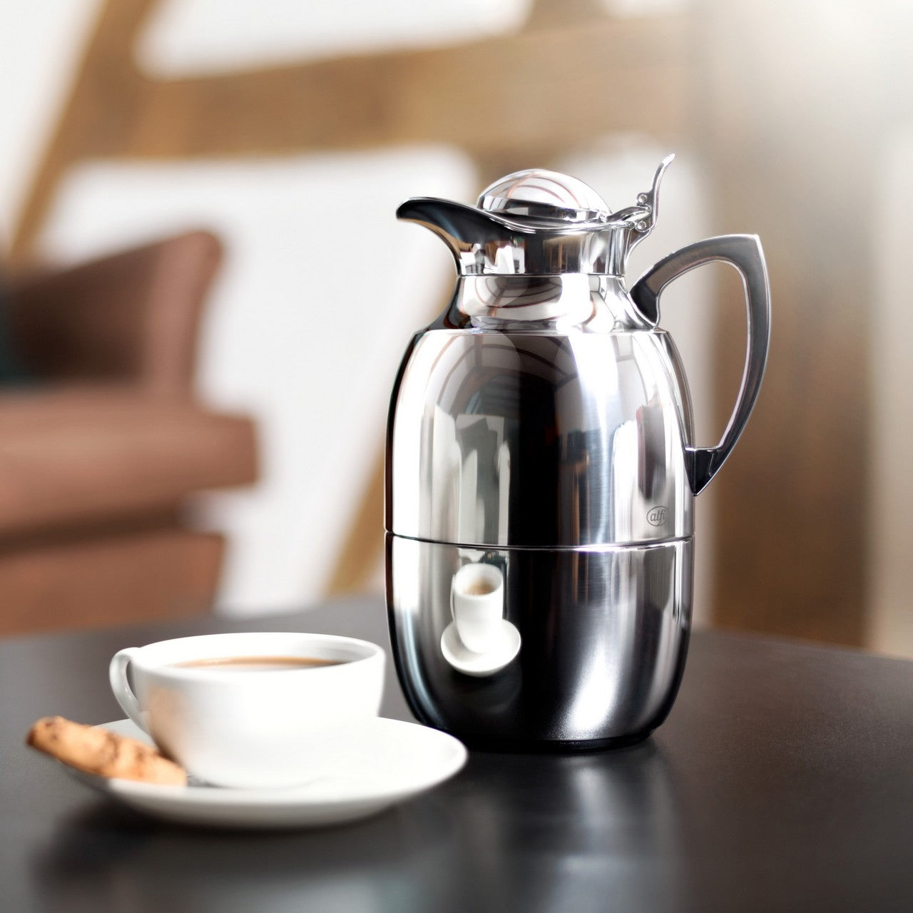 Auf dem Bild steht eine glänzende Metall-Thermoskanne mit Griff und Ausgießer. Daneben befindet sich eine weiße Tasse mit Kaffee und zwei Keksen auf einer Untertasse. Im Hintergrund ist unscharf ein Möbelstück zu sehen.