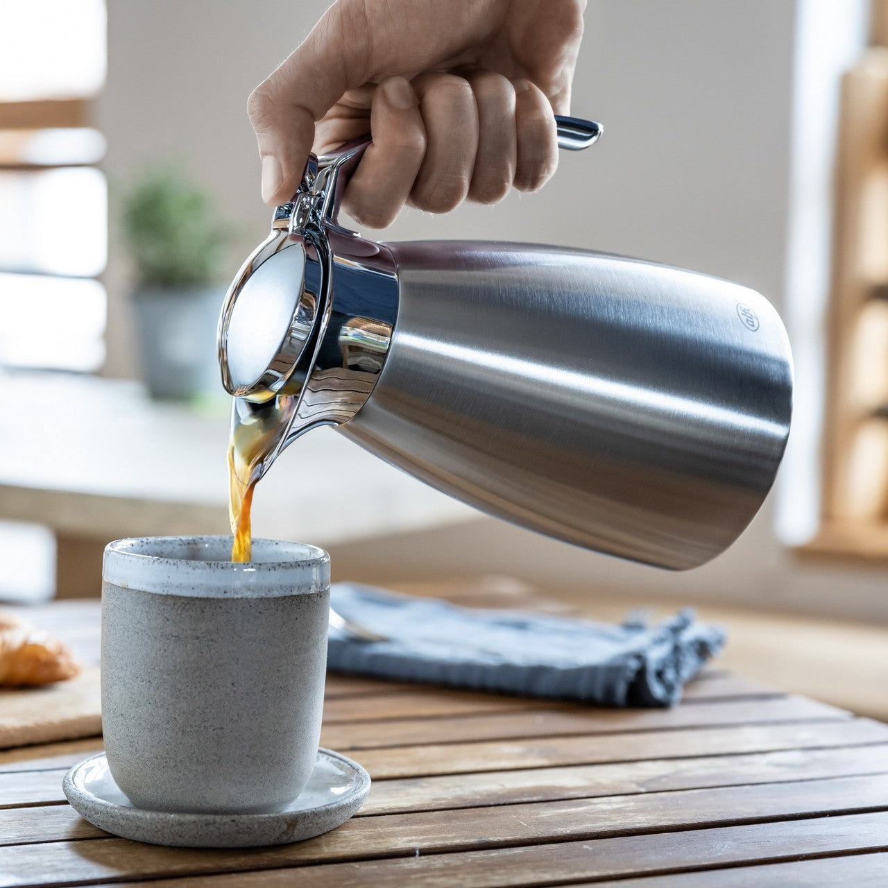 Eine Hand hält eine Kaffeekanne und gießt Orangensaft in eine weiße Tasse. Der Hintergrund zeigt einen Tisch mit einem Buch und Pflanzen in einem hellen Raum.