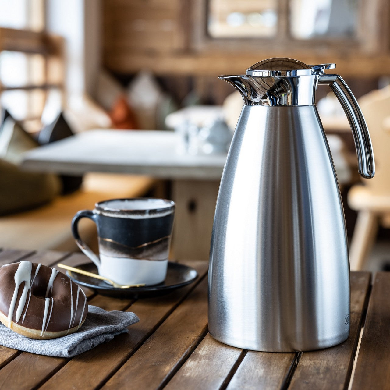 Auf einem Holztisch steht eine glänzende Thermoskanne aus Metall. Daneben befinden sich ein Glas mit Löffel und einige Schokoladenkekse auf einem Teller. Der Hintergrund zeigt unscharf ein gemütliches, warm eingerichtetes Café oder Wohnzimmer.