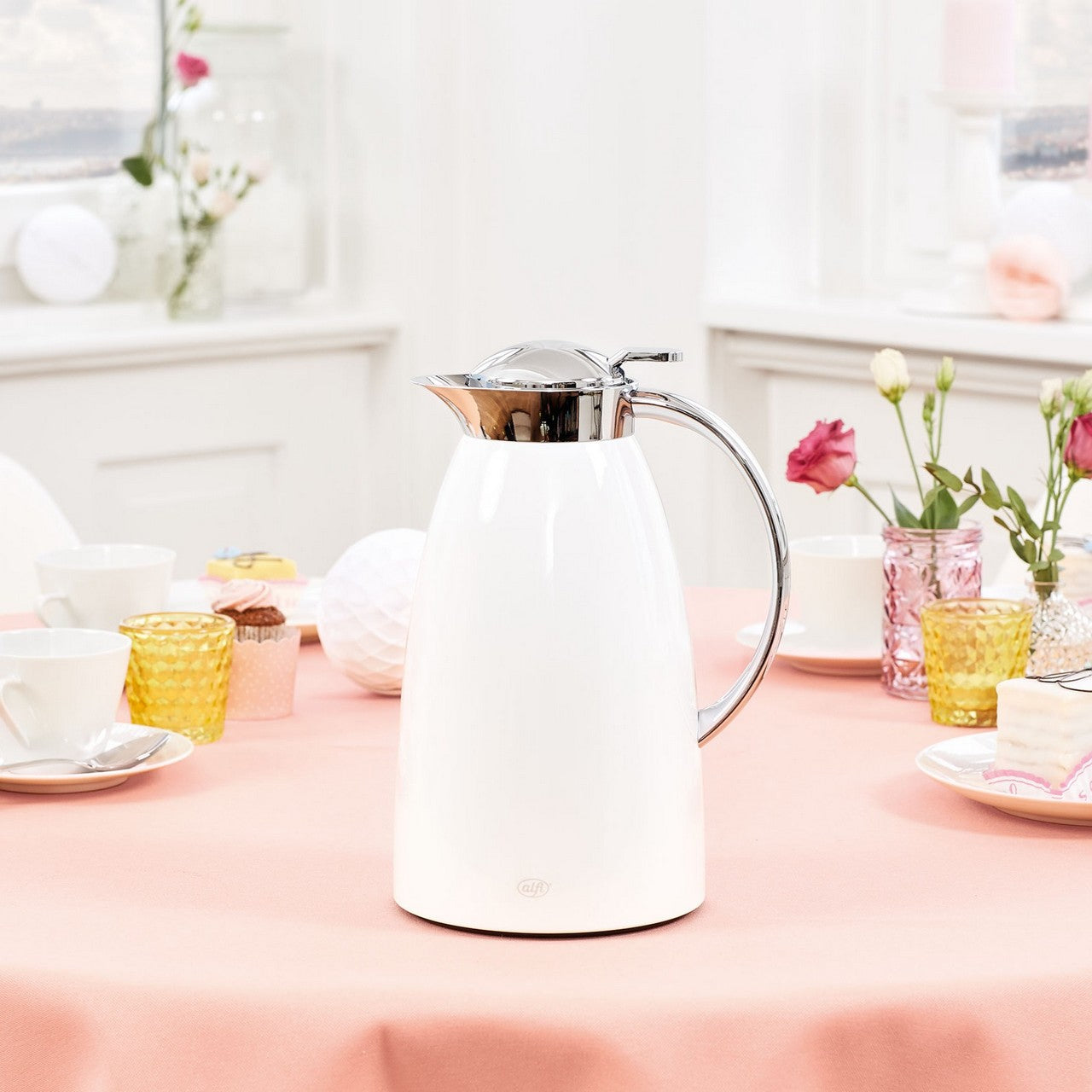 Die Szene zeigt eine weiße Kanne mit Metallgriff auf einem Tisch mit rot-weißem Tischtuch. Im Hintergrund sind unscharf Gläser mit orangefarbenen Getränken und Blumen zu sehen. Die Atmosphäre wirkt hell und freundlich.