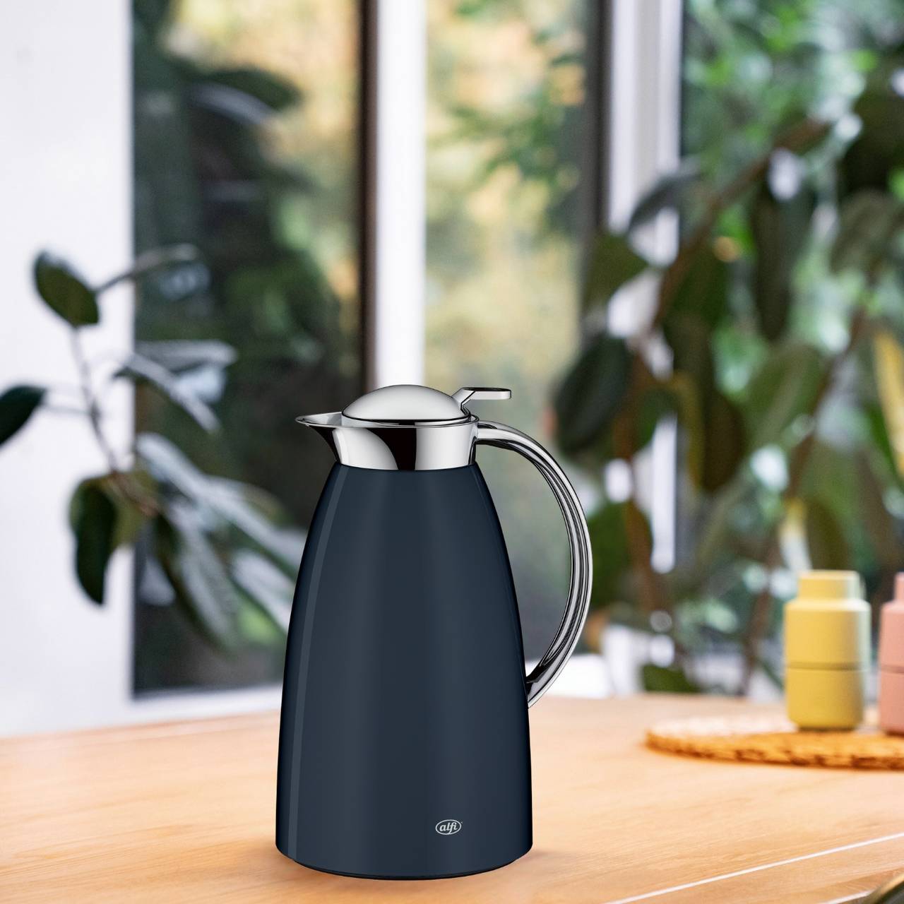Auf einem Holztisch steht eine schlanke, dunkelblaue Thermoskanne mit einem silbernen Deckel und Griff. Im Hintergrund sind unscharfe Pflanzen und große Fenster zu sehen, die auf eine grüne Umgebung hindeuten.