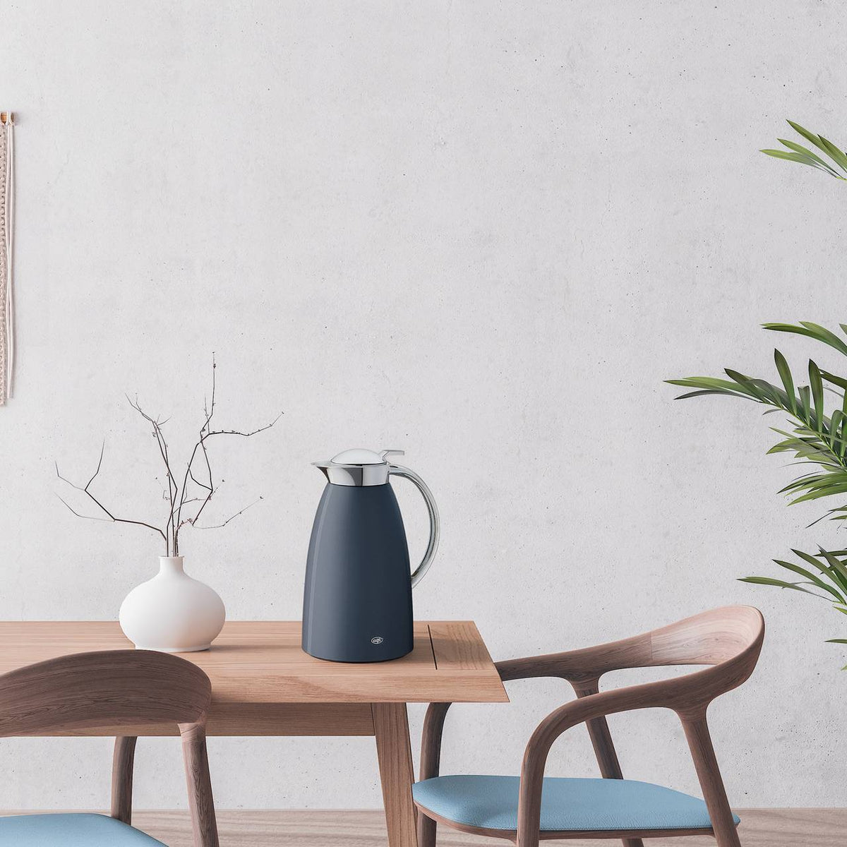 In einem modernen Raum steht ein Holztisch mit einer dunklen Thermoskanne und einer weißen Vase mit kahlen Zweigen. Im Hintergrund ist eine weiße Wand, rechts steht eine grüne Pflanze. Zwei blaue Stühle mit Holzrahmen umrahmen den Tisch.