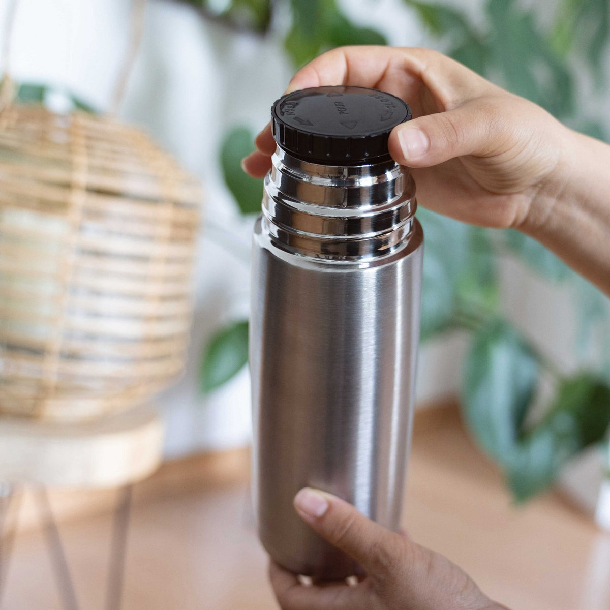 Eine Hand hält eine silberne Thermosflasche, während die andere Hand den schwarzen Schraubverschluss öffnet. Im Hintergrund sind grüne Pflanzen und ein Teil eines Lampenschirms zu sehen.