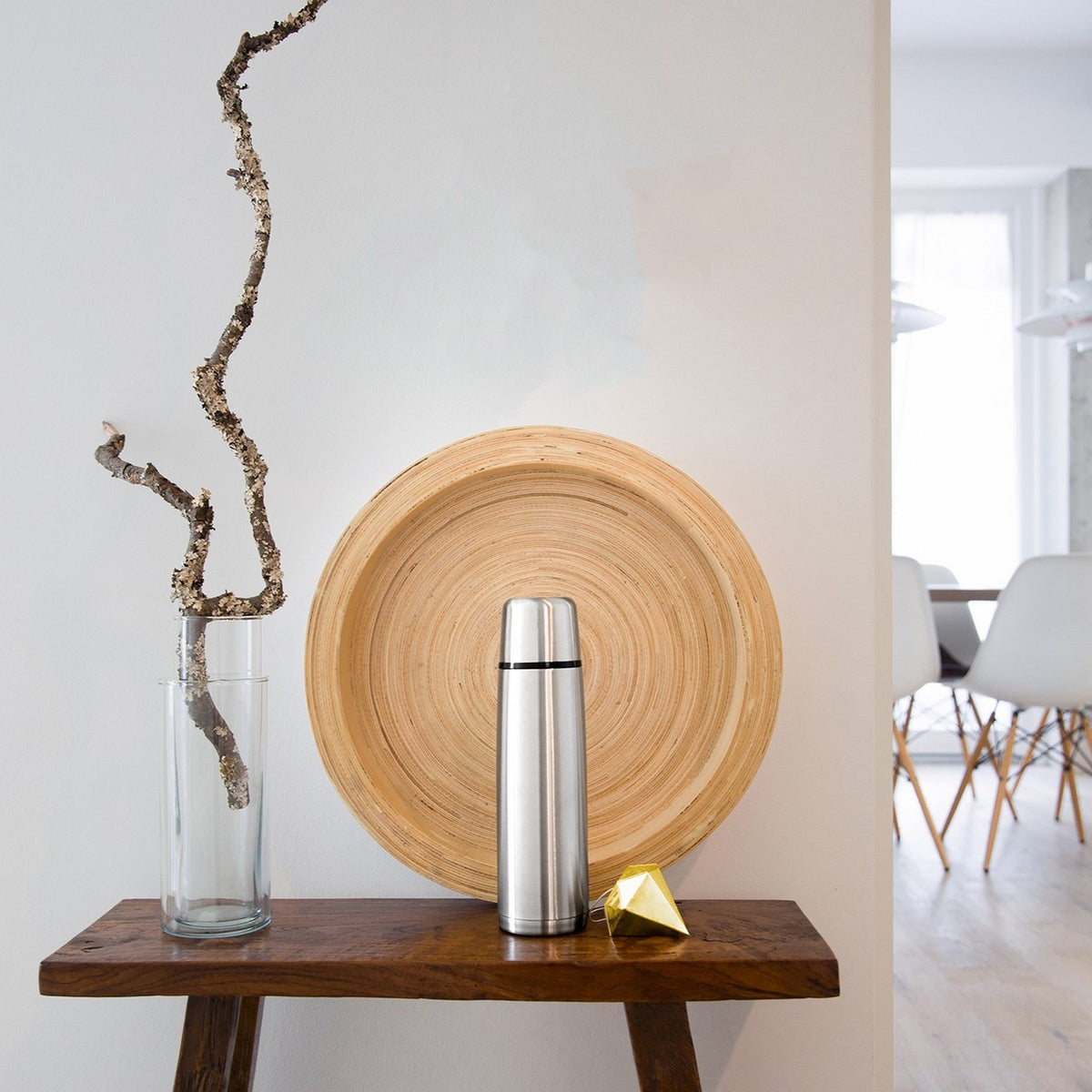 Auf einem Tisch stehen ein Glas mit einem Zweig und eine silberne Thermoskanne vor einem Holztablett. Daneben liegt ein kleines, goldfarbenes geometrisches Objekt. Im Hintergrund sind unscharf weiße Stühle erkennbar.