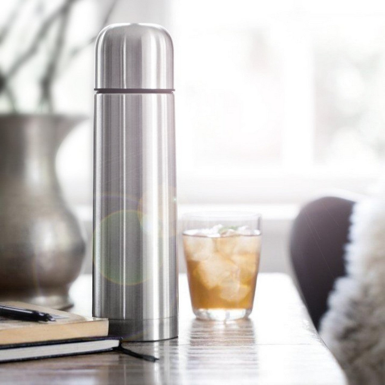 Das Bild zeigt einen silbernen Thermosbecher auf einem Tisch, daneben steht eine Vase mit Pflanzen. Im Hintergrund ist verschwommen ein Fenster mit einfallendem Licht zu sehen.