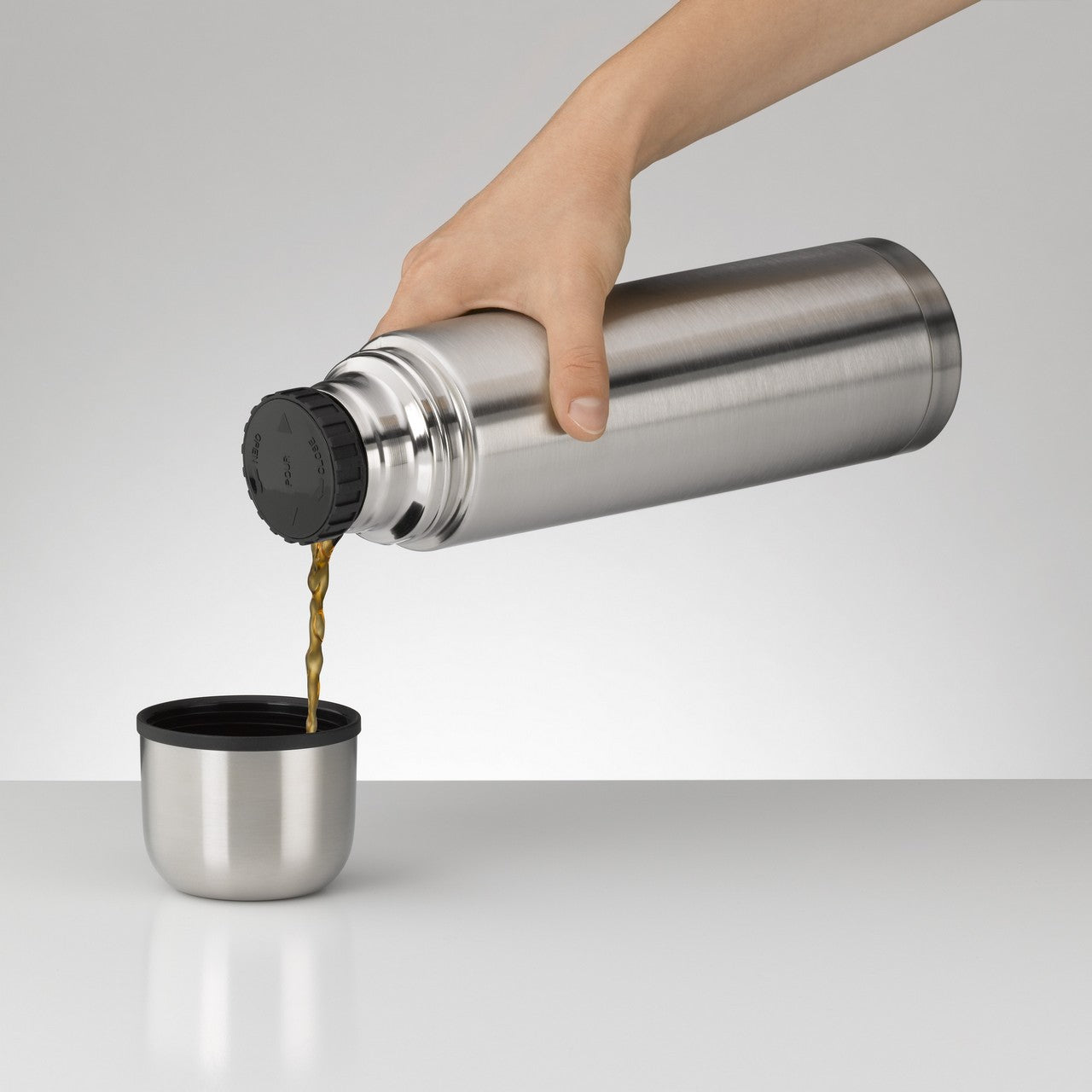 Eine Hand hält eine silberne Thermoskanne, aus der eine Flüssigkeit in eine kleine, passende Tasse gegossen wird. Der Hintergrund ist schlicht und der Fokus liegt auf der Kanne und der Tasse.