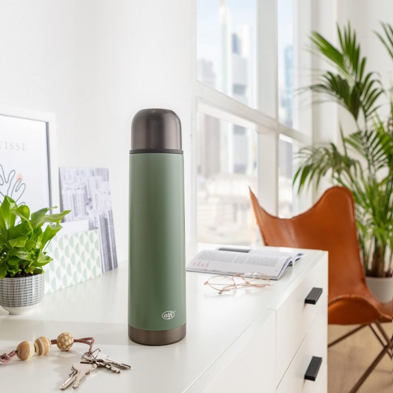 Ein moderner, heller Raum zeigt im Vordergrund eine grüne Thermoskanne auf einem weißen Tisch. Im Hintergrund gibt es einen Holzstuhl, ein großes Fenster mit viel Tageslicht und grüne Pflanzen.