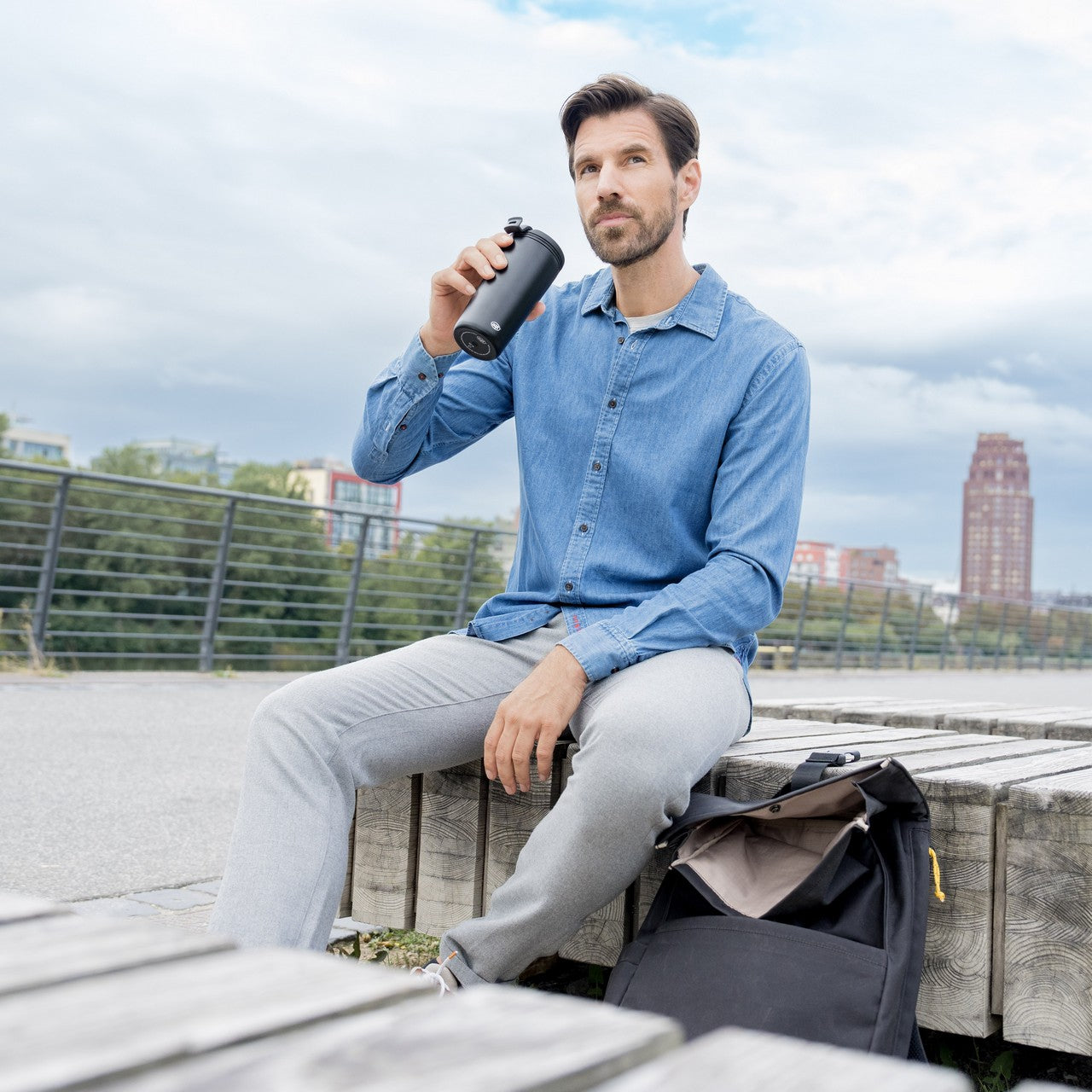 Ein Mann sitzt entspannt auf einem hölzernen Steg am Wasser und trinkt aus einer Wasserflasche. Hinter ihm sind Bäume und moderne Gebäude zu sehen. Neben ihm liegt eine schwarze Tasche.