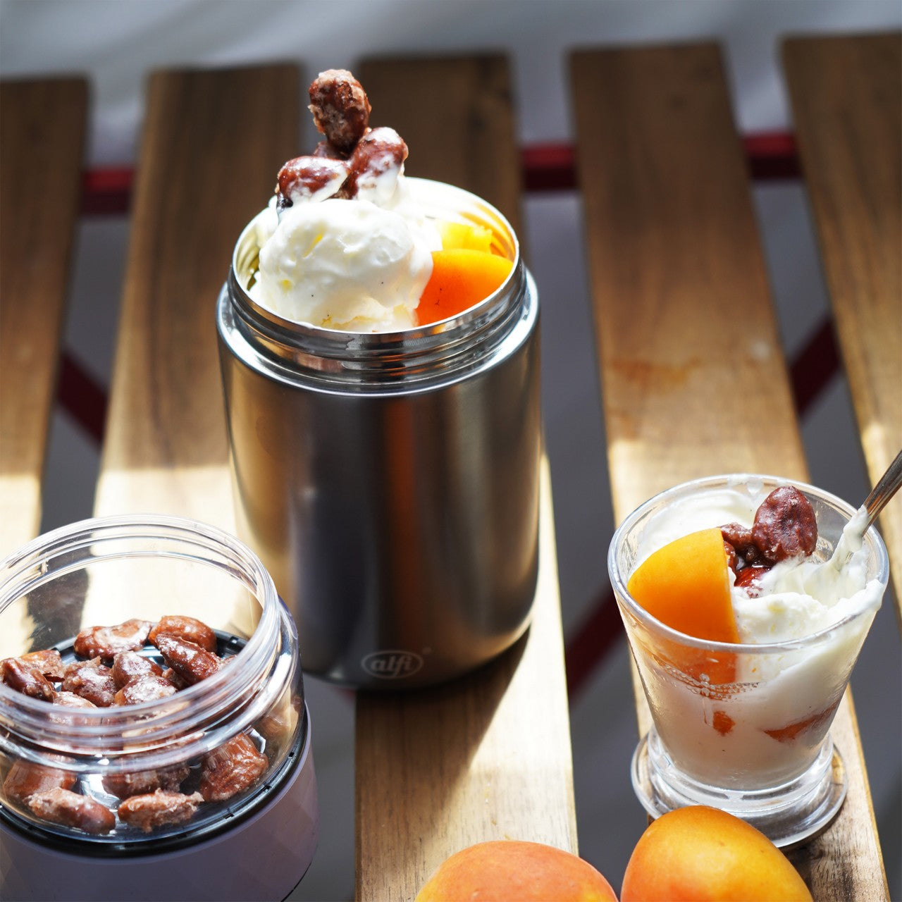 Auf einem Holztisch stehen mehrere Gefäße: Ein Edelstahlbecher mit Joghurt, frischen Aprikosen und karamellisierten Nüssen, ein Glas mit ähnlichem Inhalt und ein weiteres Glas mit Nüssen. Vor den Gefäßen liegen zwei frische Aprikosen.