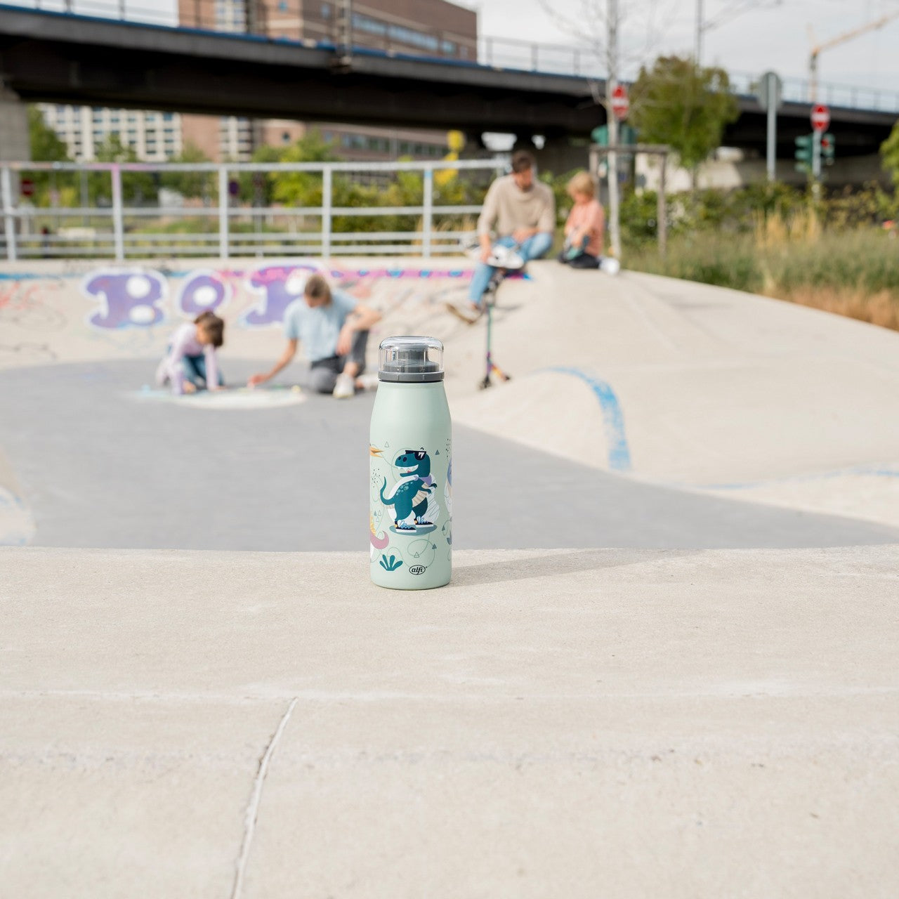 Im Vordergrund steht eine hellgrüne Trinkflasche mit Dinosaurier-Motiven. Im Hintergrund, in einem Skatepark, sitzen und spielen vier Personen auf dem Boden. Eine Brücke und einige Gebäude sind im Hintergrund zu sehen.