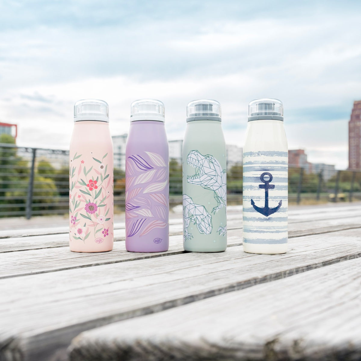 Das Bild zeigt vier stilvolle Trinkflaschen mit floralen, blätterartigen, geometrischen und nautischen Designs auf einer Holzfläche. Im Hintergrund verschwimmt eine urbane Landschaft mit Gebäuden und Himmel.