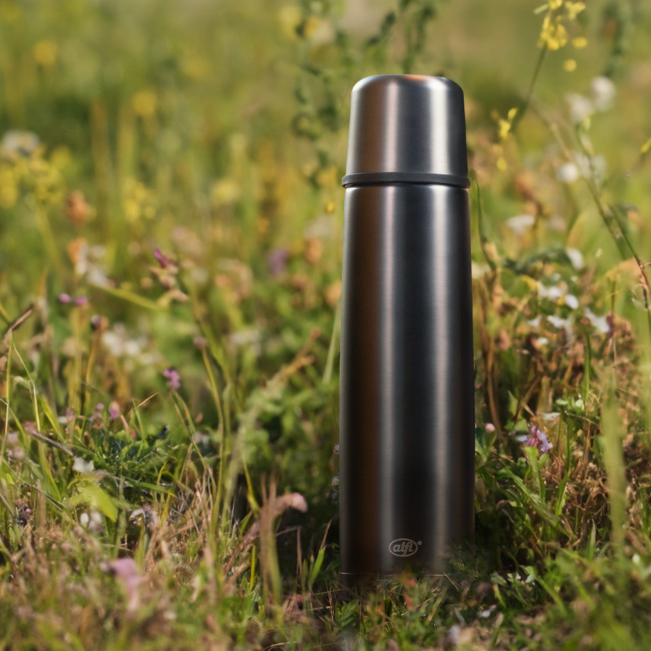 Eine Edelstahl-Thermosflasche steht in einem blühenden, wilden Blumenfeld. Die Flasche hebt sich mit ihrem schlichten, modernen Design von den bunten Blumen und grünen Pflanzen im Hintergrund ab.
