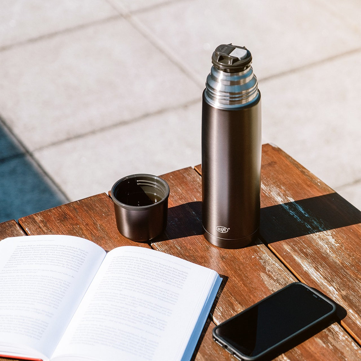 Ein geöffnetes Buch, ein Smartphone und eine Thermoskanne stehen auf einem Holztisch im Freien. Der abgenommene Deckel der Kanne dient als Becher. Sonnenlicht erzeugt deutliche Schatten.