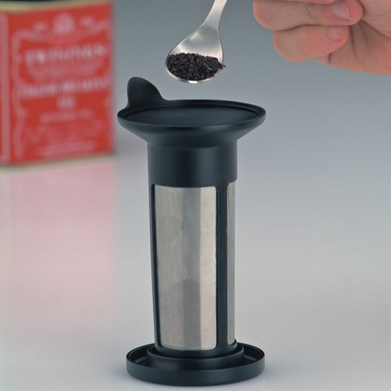 Das Bild zeigt einen kleinen, zylinderförmigen Kaffeezubereiter mit einem metallischen Filter in der Mitte. Eine Hand hält einen Löffel mit Kaffeepulver über dem Gerät. Im Hintergrund ist eine rote Verpackung zu sehen.