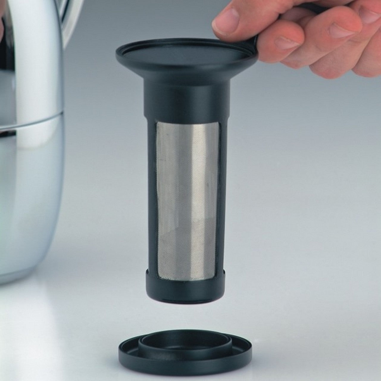 Eine Hand hält einen zylinderförmigen Edelstahlfilter mit schwarzem Griff über einer schwarzen Unterlage. Links ist ein Teil einer silbernen Kanne sichtbar. Der Filter passt wohl in die Kanne.