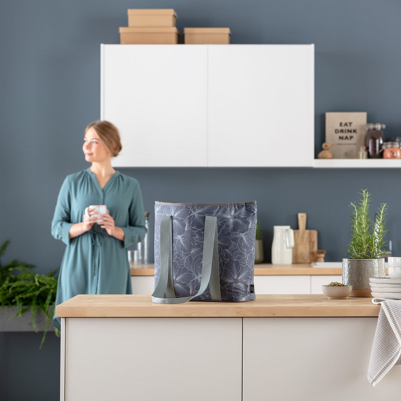 In einer modernen Küche mit weißen Schränken hält eine Frau in einem blauen Kleid einen Becher. Auf der Kücheninsel steht eine gemusterte Tragetasche. Dekorative Pflanzen sorgen für ein ruhiges Ambiente.