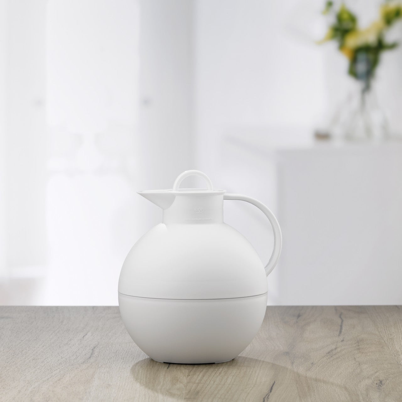 Das Bild zeigt eine runde, weiße Thermoskanne mit einem Henkel und einem kleinen Ausguss, die auf einem Holztisch steht. Der Hintergrund ist verschwommen und zeigt eine helle, minimalistische Einrichtung mit Blumen.