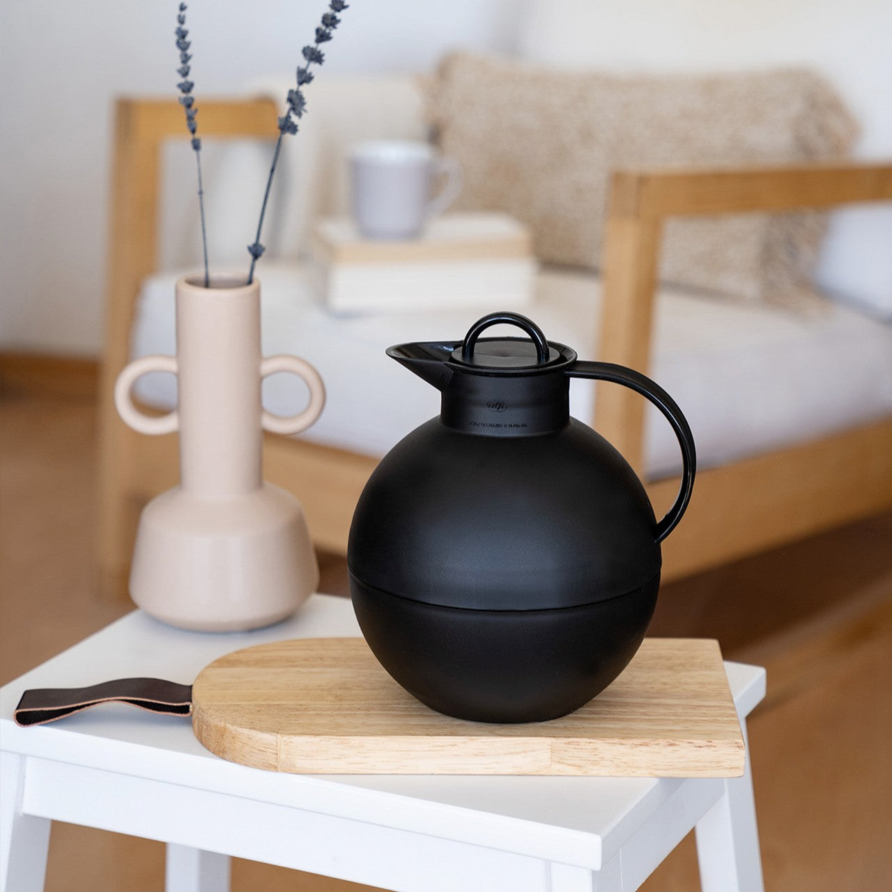 Auf einem Holzbrett steht eine schwarze, kugelförmige Kanne. Links daneben befindet sich eine beige Vase mit Lavendel. Im Hintergrund sieht man ein helles Sofa mit einem Kissen.