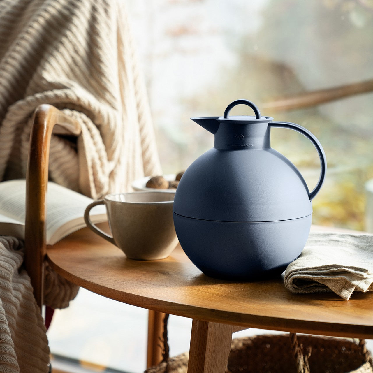 Auf einem Holztisch stehen eine runde, blaue Thermoskanne und eine graue Tasse. Daneben liegen ein aufgeschlagenes Buch und eine beige Decke. Helles, diffuses Licht kommt durch ein unscharfes Fenster im Hintergrund.