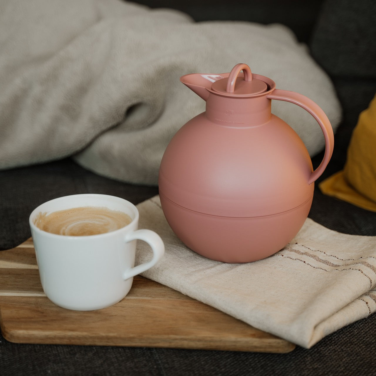 Auf dem Bild steht eine rosa Thermoskanne neben einer Tasse Kaffee mit Milchschaum auf einem Holzbrett. Das Ganze ist auf einem weichen, beigefarbenen Stoff platziert, mit einem gelben Kissen im Hintergrund für Gemütlichkeit.