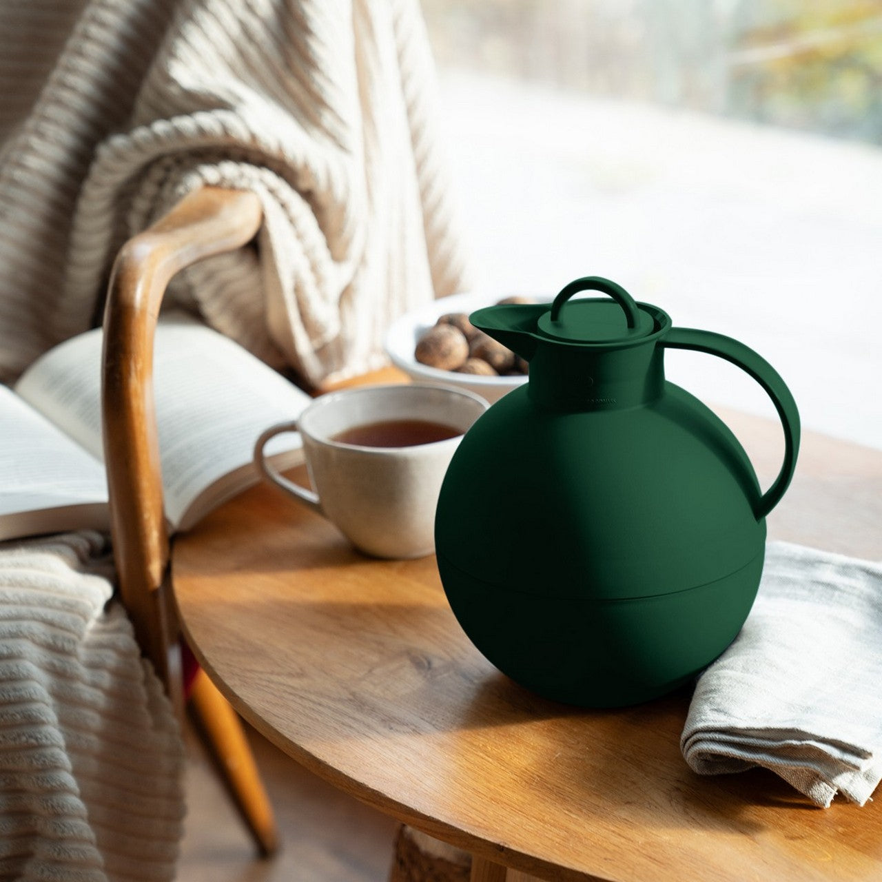 Auf einem kleinen Tisch steht eine grüne Kanne neben einer Tasse Tee. Daneben liegt ein Buch auf einem gemütlichen Sessel mit einer weichen Decke. Im Hintergrund sind Nüsse sichtbar, die auf einem Teller liegen.