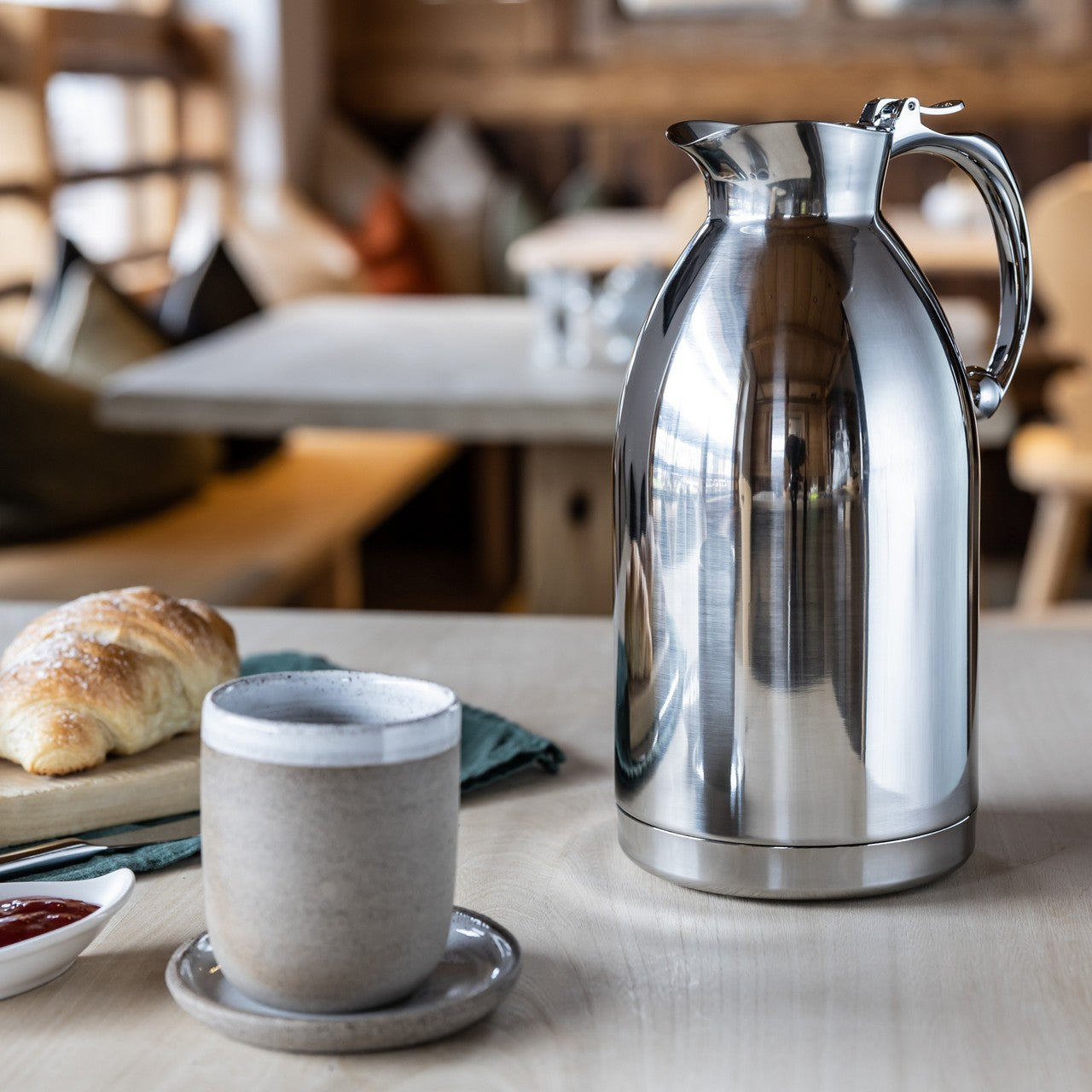 Ein silberner Isolierkrug und eine weiße Tasse stehen auf einem Tisch, daneben ein Croissant auf einem Teller. Im Hintergrund sind unscharf Möbel zu erkennen, die auf eine gemütliche Café-Atmosphäre schließen lassen.