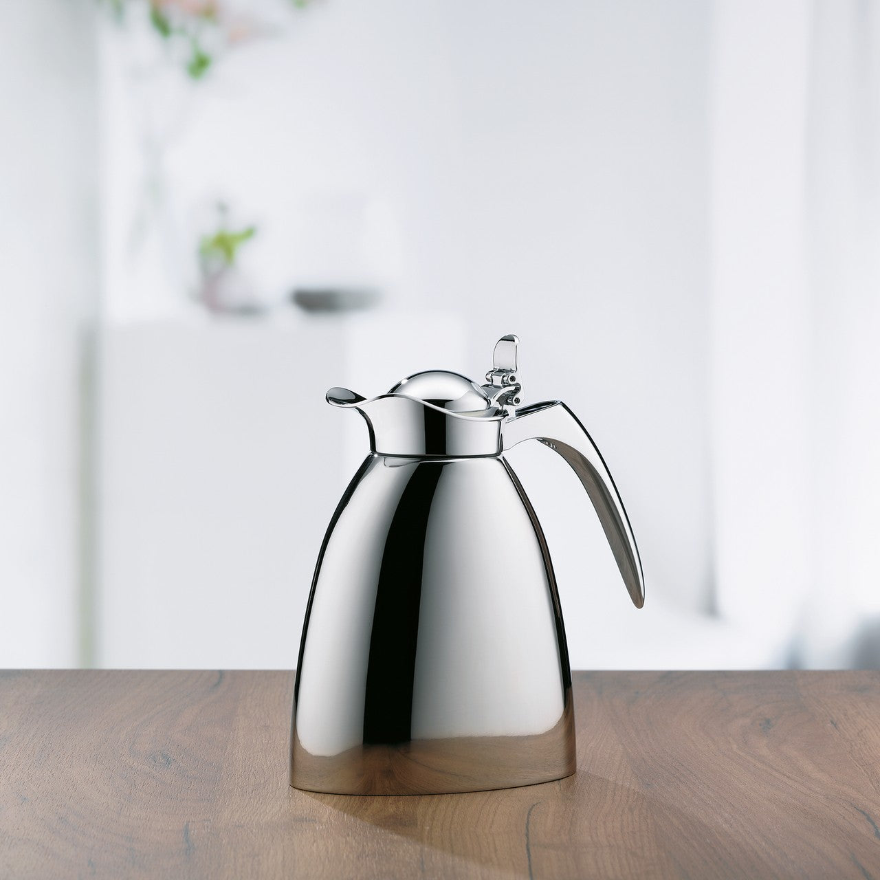 Das Bild zeigt eine glänzende, silberne Isolierkanne mit elegant geschwungenem Griff, die auf einer Holzoberfläche steht. Im Hintergrund ist ein unscharfer, heller Raum mit Blumen und Dekorationsobjekten zu sehen.