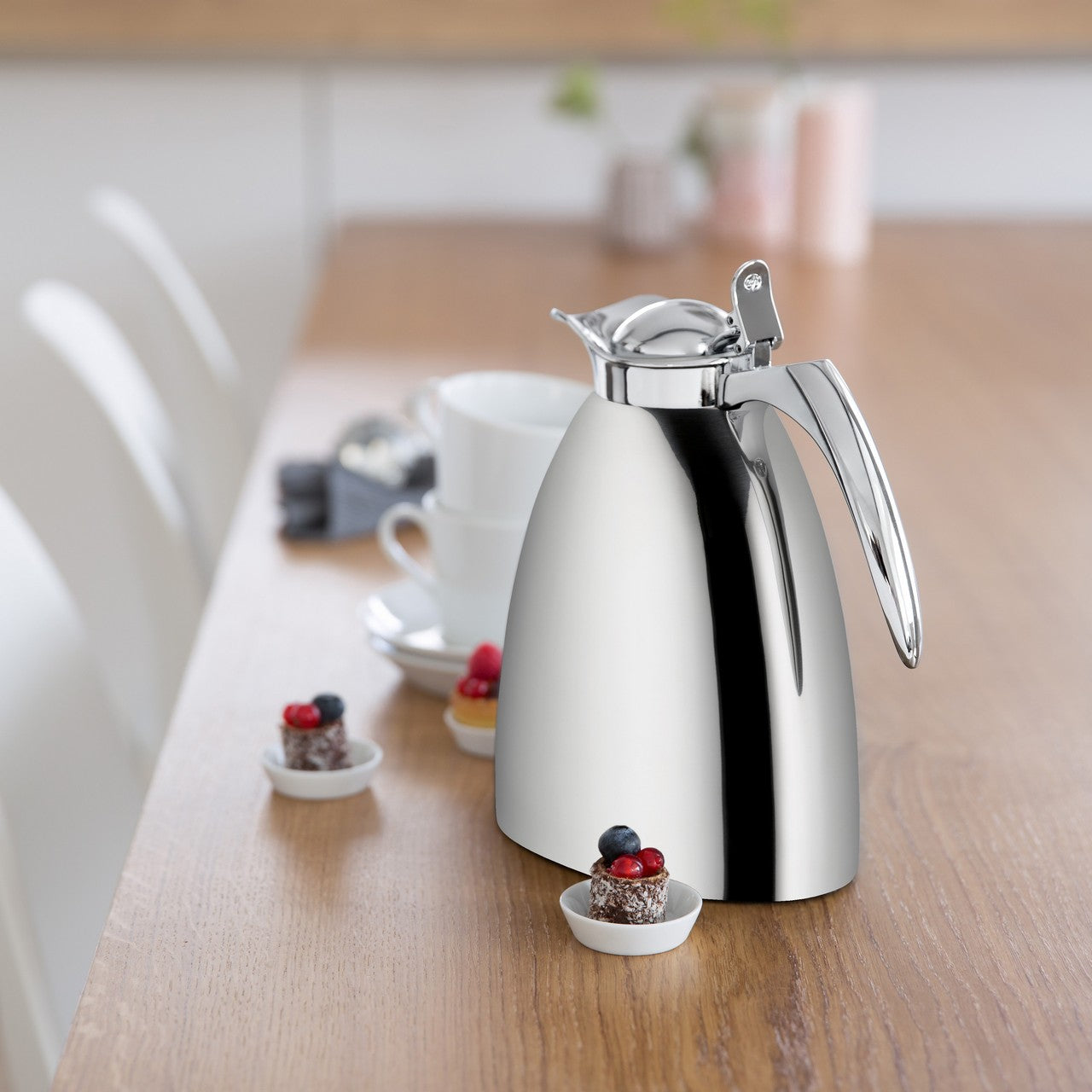 Auf dem Bild steht eine silberne Thermoskanne auf einem Holztisch. Daneben befinden sich weiße Tassen und kleine, mit Beeren garnierte Desserts in weißen Schälchen. Im Hintergrund sind verschwommene Küchenutensilien erkennbar.