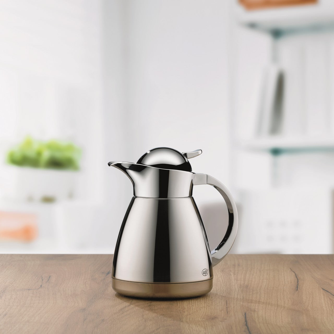 Das Bild zeigt eine glänzende, silberne Thermoskanne mit modernem Design auf einem Holztisch. Im Hintergrund sind verschwommene, weiße Regale und eine grüne Pflanze zu sehen. Die Kanne hat einen gebogenen Griff und klappbaren Ausgießer.