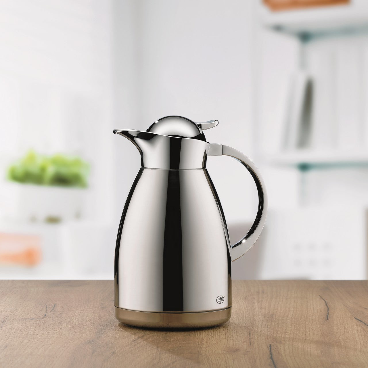 Auf dem Bild ist eine glänzende, silberne Thermoskanne auf einem Holztisch abgebildet. Sie hat einen eleganten Griff und einen gut sichtbaren Ausgießer. Im Hintergrund sind unscharf Regale und eine Pflanze zu erkennen.