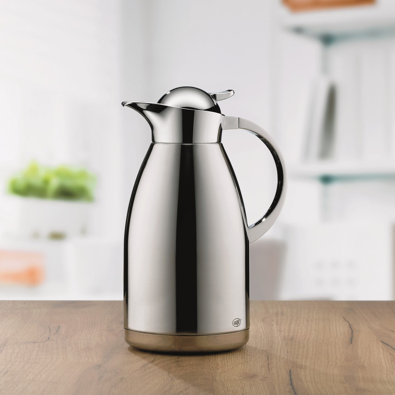 Ein glänzender, silberner Thermoskrug steht auf einem Holztisch vor einem unscharfen Hintergrund. Der Krug hat einen gebogenen Griff und einen Klappdeckel. Im Hintergrund sind helle Regale und ein Pflanzentopf erkennbar.