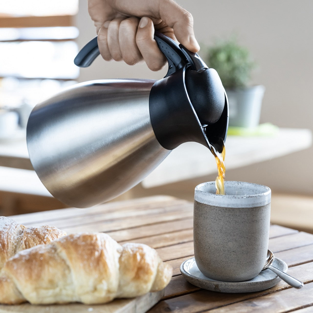 Eine Hand gießt Tee aus einer silbernen Thermoskanne in eine graue Tasse, die auf einer Untertasse mit einem kleinen Löffel steht. Auf dem Holztisch im Hintergrund liegen frische Croissants.