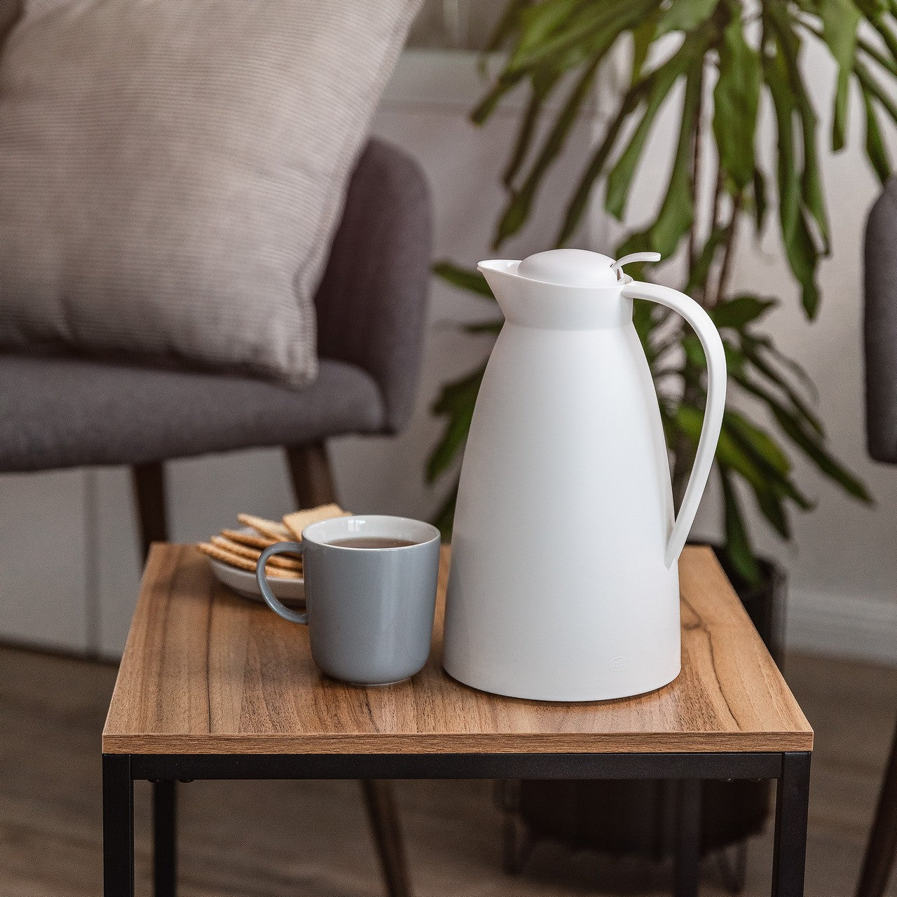 Auf einem kleinen Holztisch stehen eine weiße Kanne und eine graue Tasse mit Flüssigkeit. Daneben liegen einige Kekse auf einem Teller. Im Hintergrund ist ein gemütlicher Raum mit Pflanzen und einem Sessel zu sehen.